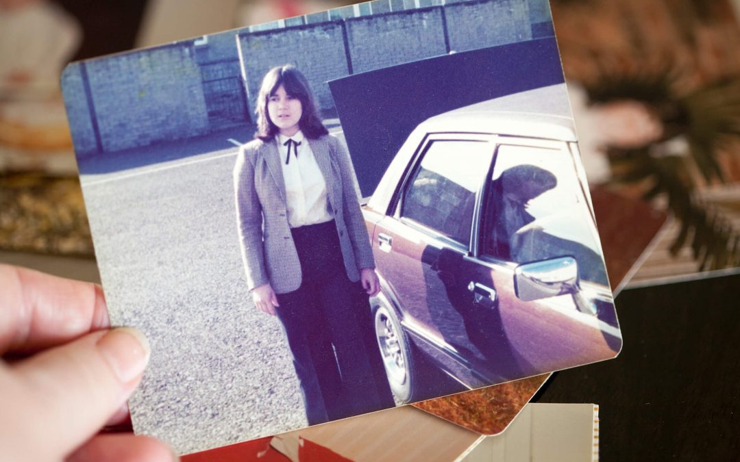 Hand holding an hold photo of a young woman standing by a car